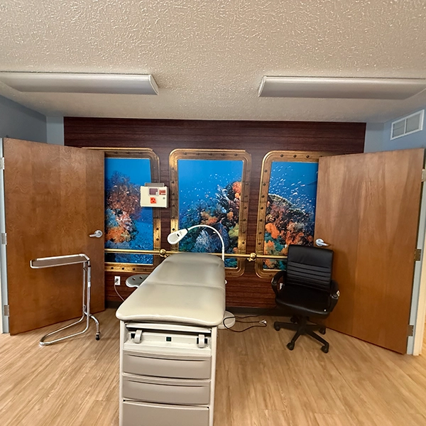 A treatment room featuring a padded exam table, a chair, and a vibrant underwater mural with coral and fish behind two wooden doors.